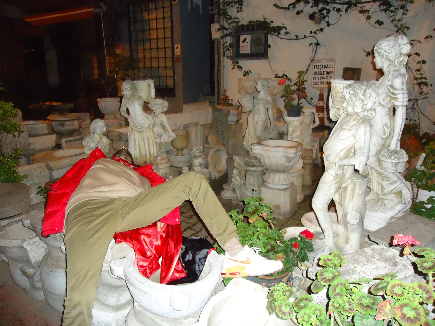 Person lying among white garden statues next to a red bag