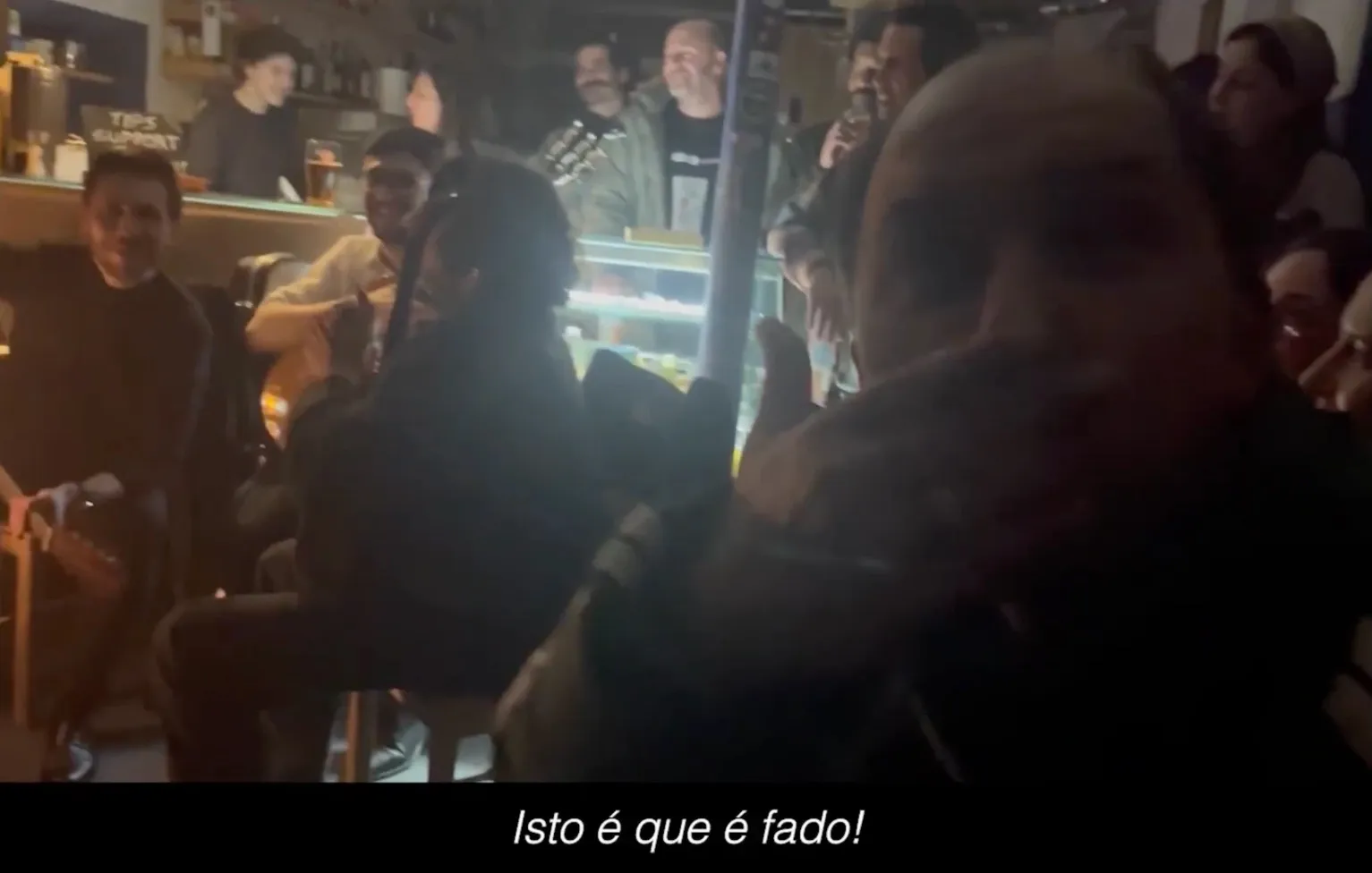 Musicians and listeners in a crowded bar, with the subtitle “Isto é que é fado!”